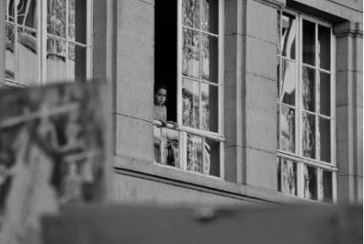 A lonely girl gazes through a window in a great house.